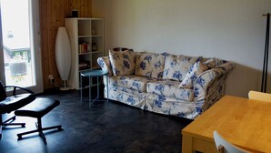 Ein Zimmer mit Holzboden, einem blumigen Sofa, einem Holztisch, einem Bücherregal und einem Fenster mit Ausblick.