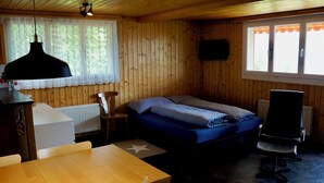 Ein Zimmer in einer Holzhütte mit einem Bett, einem Schreibtisch, einem Stuhl, einem Tisch, einem Heizkörper und einem an der Wand befestigten Fernseher.