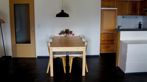 Ein Essbereich mit Holztisch und Stühlen, einer Vase mit Blumen, einer schwarzen Pendelleuchte und Holzschränken im Hintergrund.