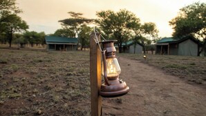 A rustic wooden post with a lantern attached, set against a backdrop of a dirt path and sparse vegetation, with distant structures that appear to be huts or cabins.