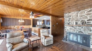 Un interior de madera con chimenea de piedra, un área de comedor con un tablero de ajedrez y una cocina al fondo.