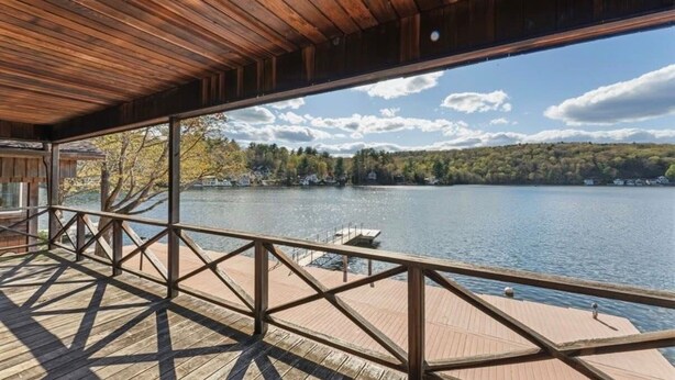 Una terraza de madera con barandilla da vista a un lago, un muelle y una orilla boscosa.
