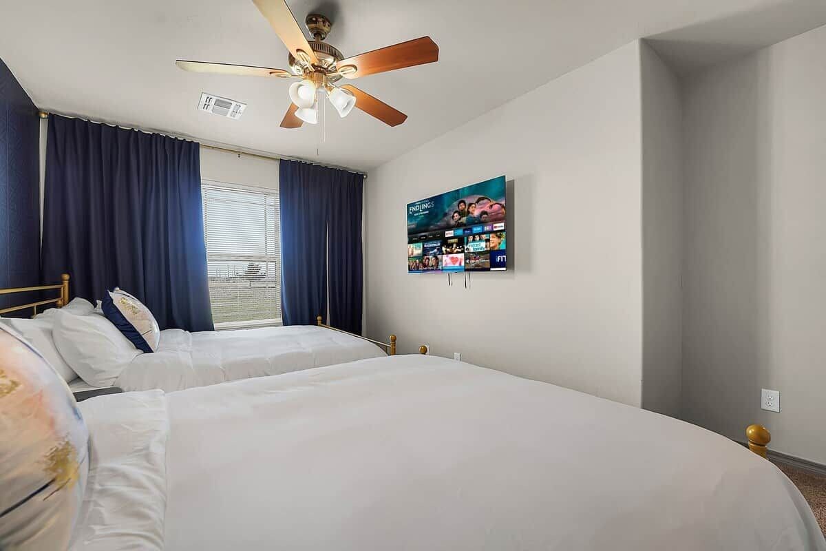 A hotel room with two beds, a ceiling fan, a large window with curtains, and a wall-mounted television.