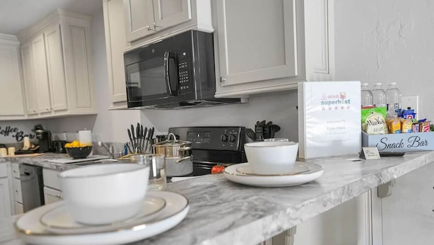 Una cocina con armarios blancos, una estufa negra y una encimera con una cafetera, utensilios y una caja etiquetada 'Super Host'.