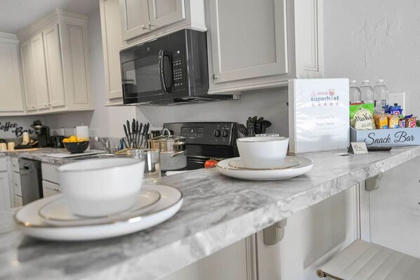 A kitchen with white cabinets, a black stove, and a countertop with a coffee machine, utensils, and a box labeled 'Super Host.'