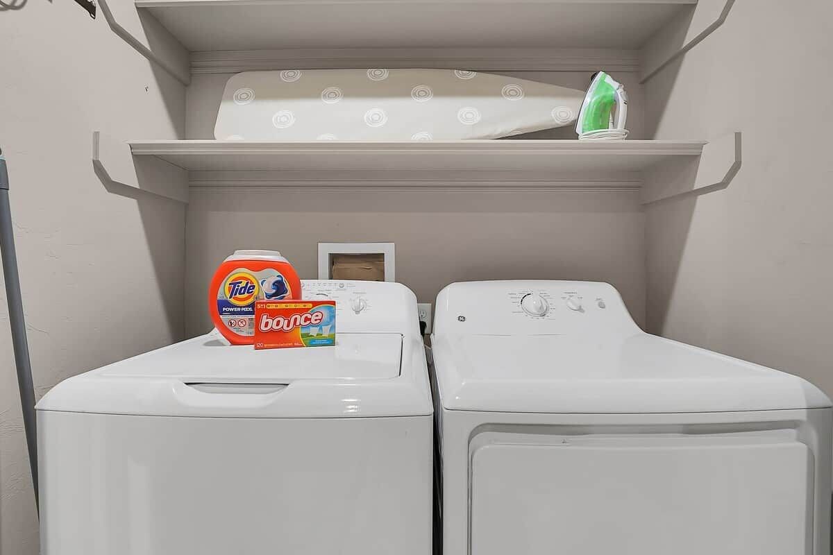 A laundry area with two white washing machines, a shelf above with a green item, and Tide Bounce detergent on the left machine.