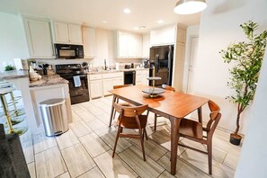 A modern kitchen with white cabinets, a stainless steel refrigerator, a wooden dining table with chairs, and a potted plant.