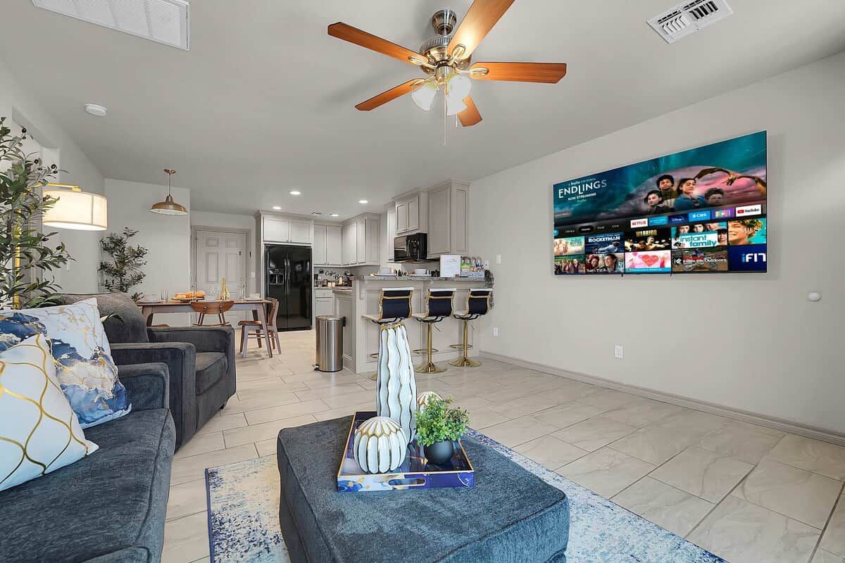 A modern living room with a sofa, a coffee table with decorative items, a ceiling fan, and a wall-mounted TV displaying a movie poster.