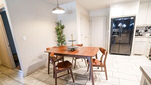 Un coin repas avec une table et des chaises en bois, un luminaire suspendu, une plante en pot et, au fond, un réfrigérateur en acier inoxydable.
