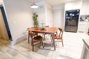 A dining area with a wooden table and chairs, a hanging light fixture, a potted plant, and a stainless steel refrigerator in the background.