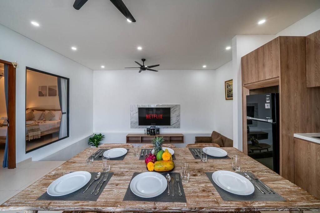Uma sala de jantar moderna com uma mesa de madeira posta para quatro pessoas, um ventilador de teto e uma TV exibindo 'NETFLIX'.