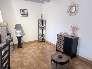 A room with a tiled floor, a round table, a black cabinet, a lamp, a bookshelf, and a wall-mounted mirror. - Ty Ar Méné Vacation Rental (Lohuec)