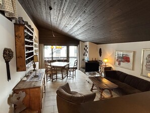 Een gezellige woonkamer met houten balkenplafond, een zithoek met tafel en stoelen, en een open haard.