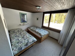 A small, single-room accommodation with two beds, a ceiling light fixture, a window with curtains, and a framed picture on the wall.