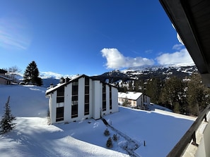 Schneebedeckte Landschaft mit einem modernen Gebäude, das große Fenster und ein geneigtes Dach besitzt, umgeben von Bäumen und Bergen im Hintergrund.