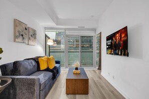 A modern living room with a grey sofa, a wooden coffee table, and a large window with a view of buildings. - Midtown Vale Hotel (Toronto)