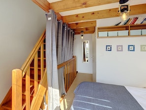Een slaapkamer met een houten trap, een bed met een grijze deken, een boekenplank vol boeken en een klein raam.