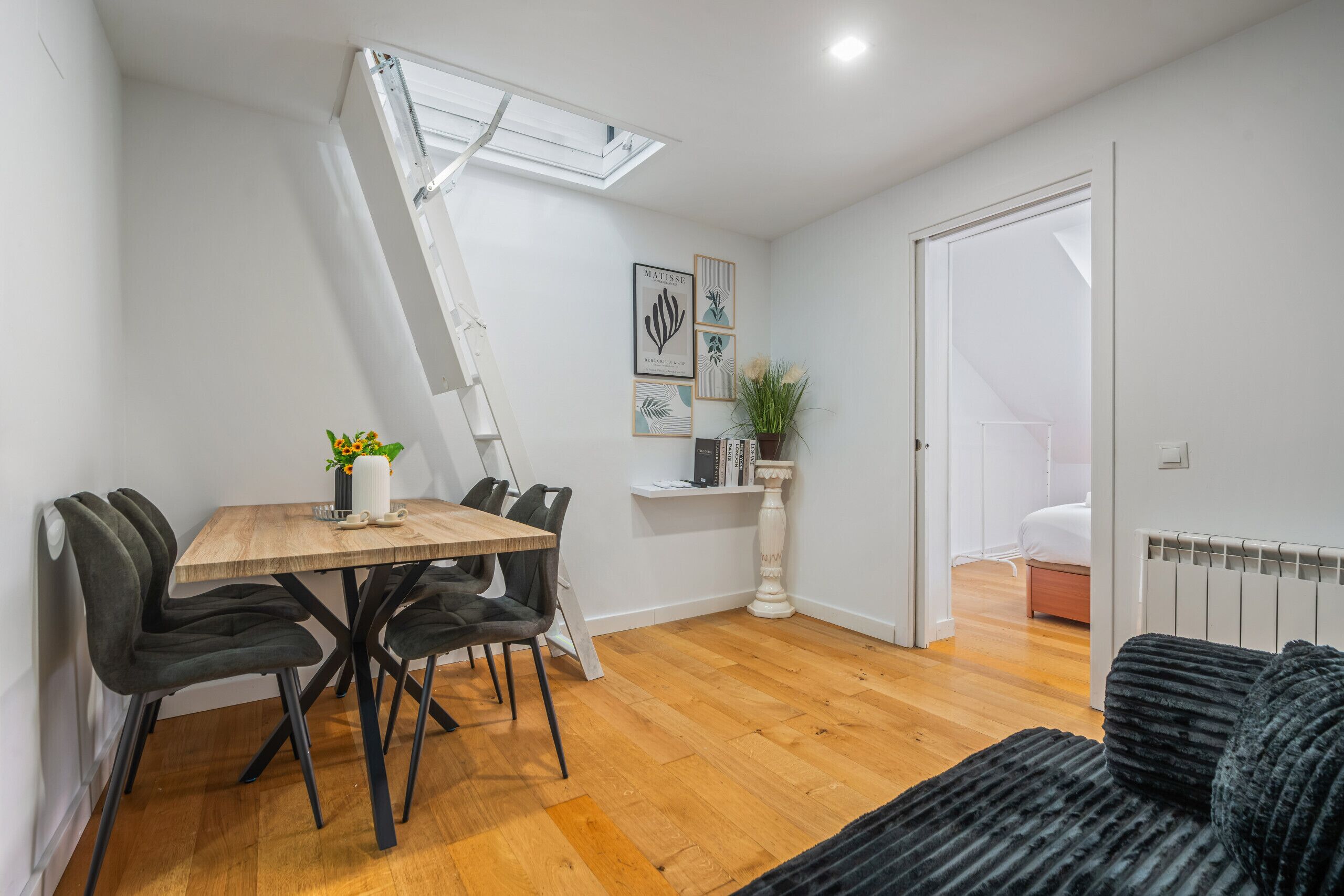 Un comedor moderno con una mesa de madera y sillas negras, un radiador blanco y vista a un dormitorio por una puerta abierta.