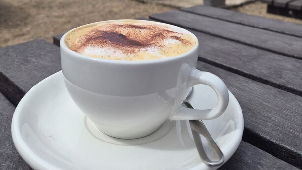 Una taza de café blanca sobre un platito blanco con una cuchara, colocada sobre una mesa de madera.