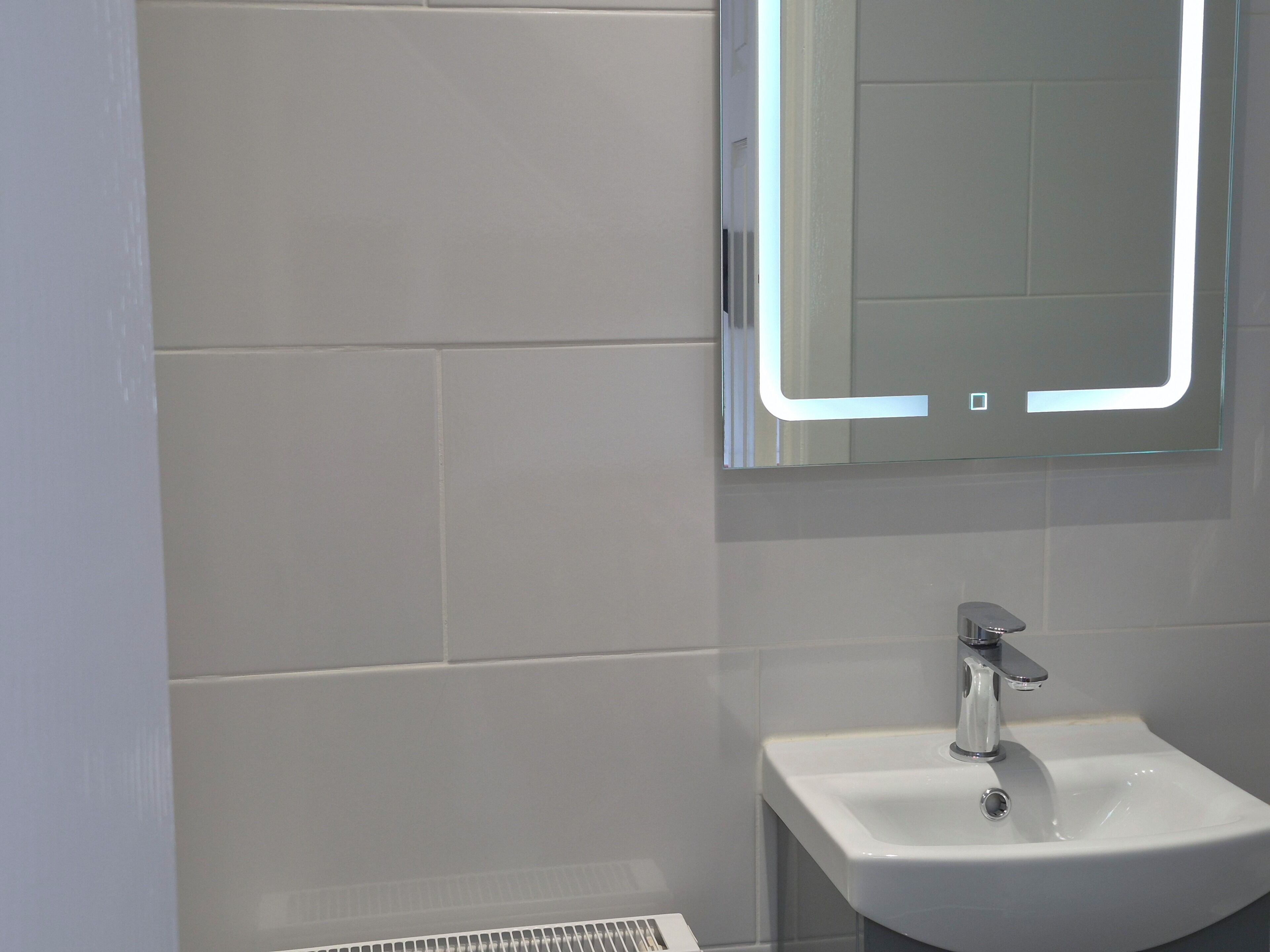 A bathroom with a white sink, a mirror with a light, and a vent on the wall.