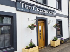 Um edifício com uma placa indicando 'The Crosswater Restaurant' e 'PUB'. Há vasos de madeira com flores na entrada.