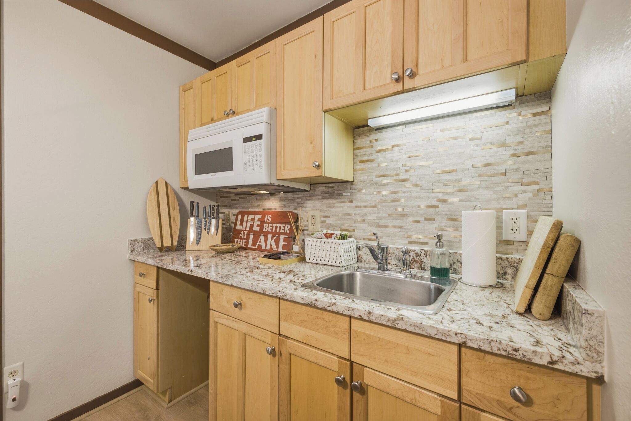 A kitchen with wooden cabinets, a granite countertop, a stainless steel sink, a microwave, and a sign that reads 'Life is better at the lake.'