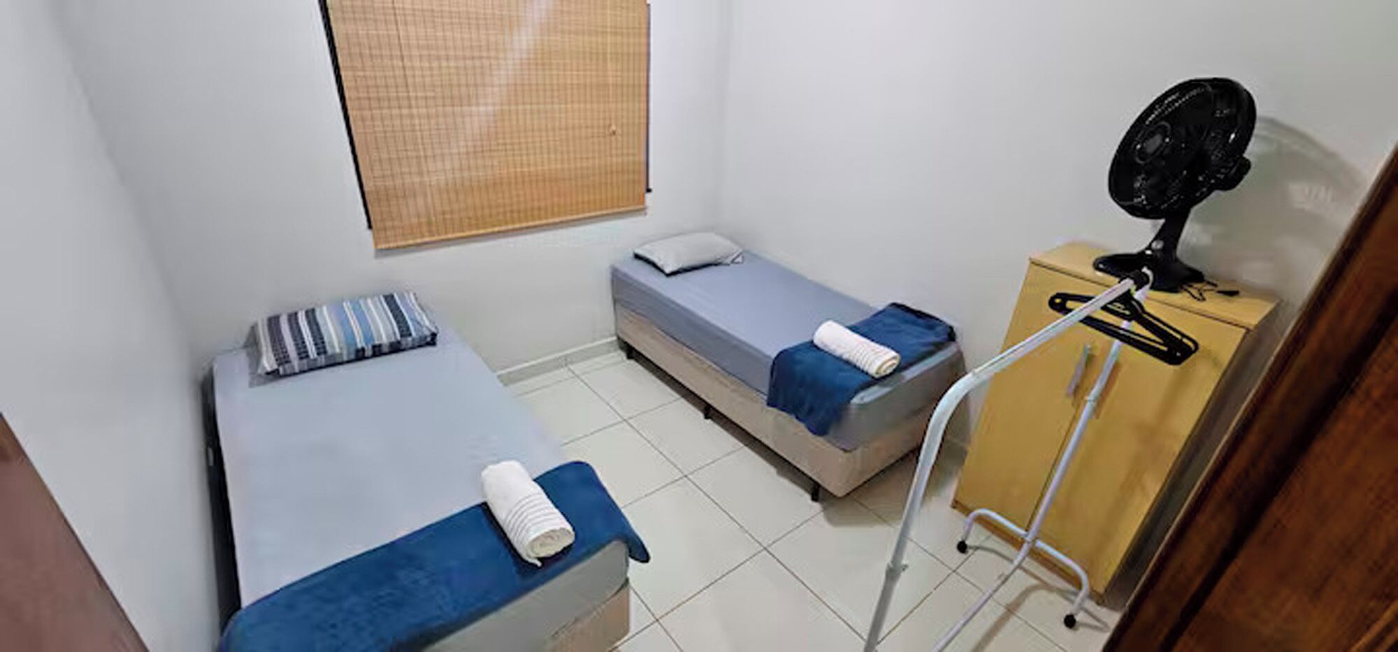 Une petite chambre propre, avec deux lits simples, une table de chevet équipée d’un ventilateur et une fenêtre avec un store.