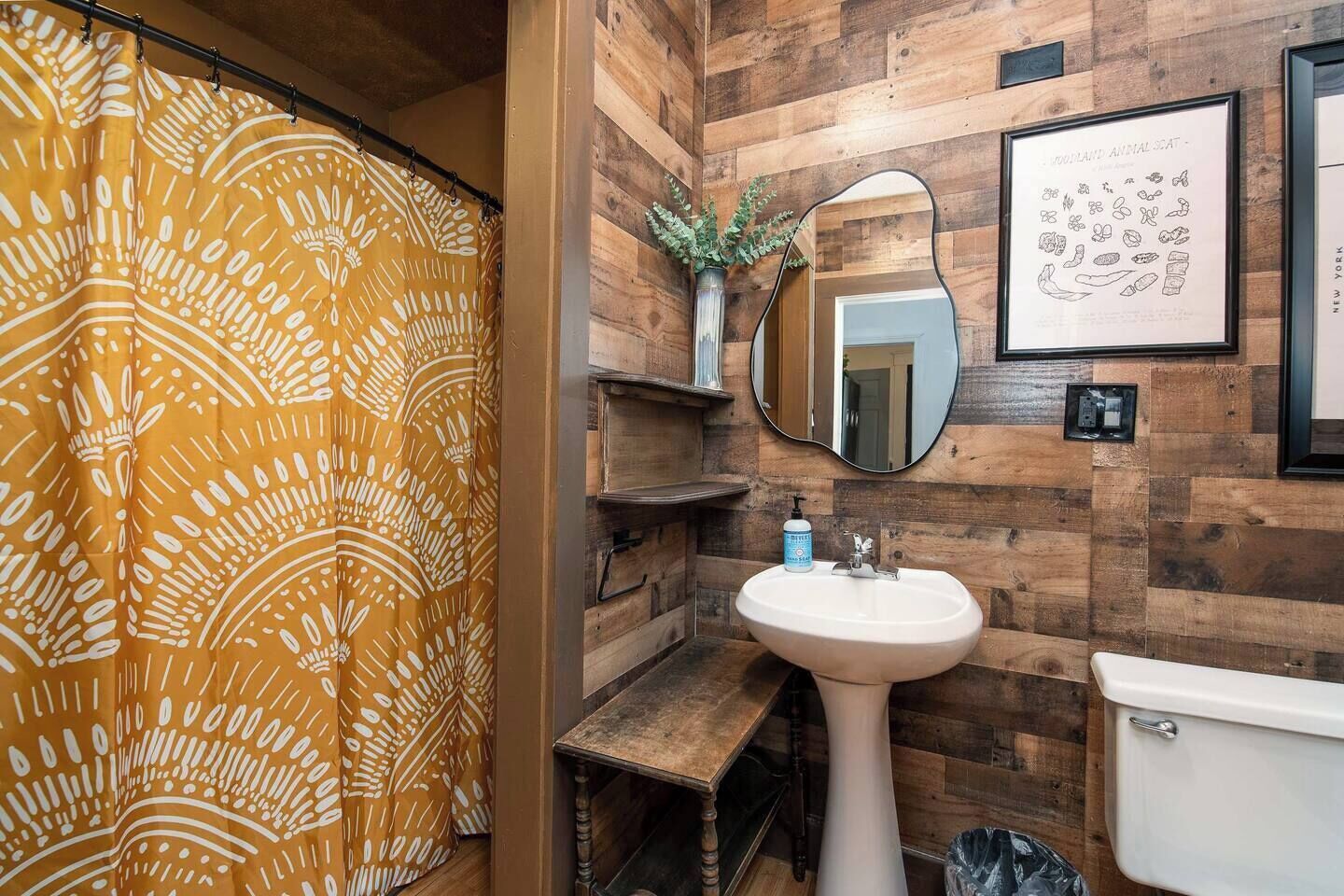 Un baño con pared de madera, lavamanos blanco, inodoro, espejo y cortina de ducha.