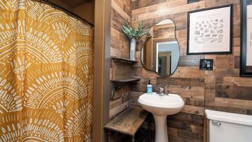 Un baño con pared de madera, lavamanos blanco, inodoro, espejo y cortina de ducha.