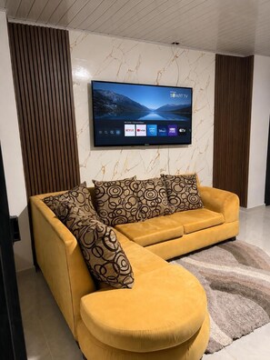 A modern living room with a yellow sofa, patterned cushions, a mounted flat-screen TV, and a marble-patterned wall.