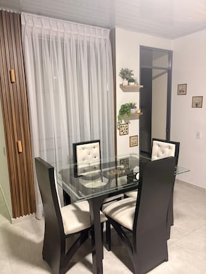 A dining area with a glass table and black chairs, a wooden panel wall, and a white curtain.