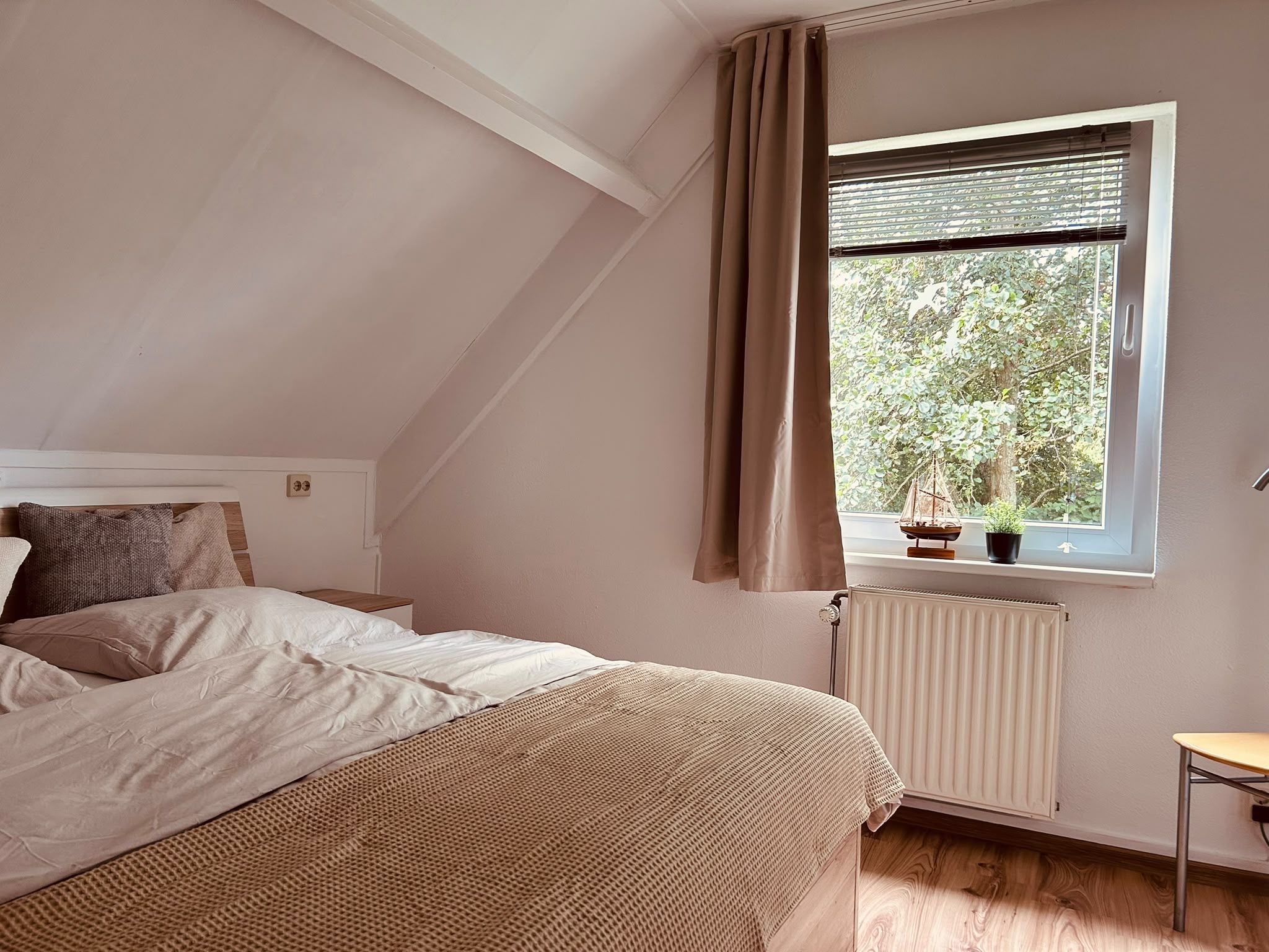 A bedroom with a bed, a window with curtains, a radiator, and a small table.