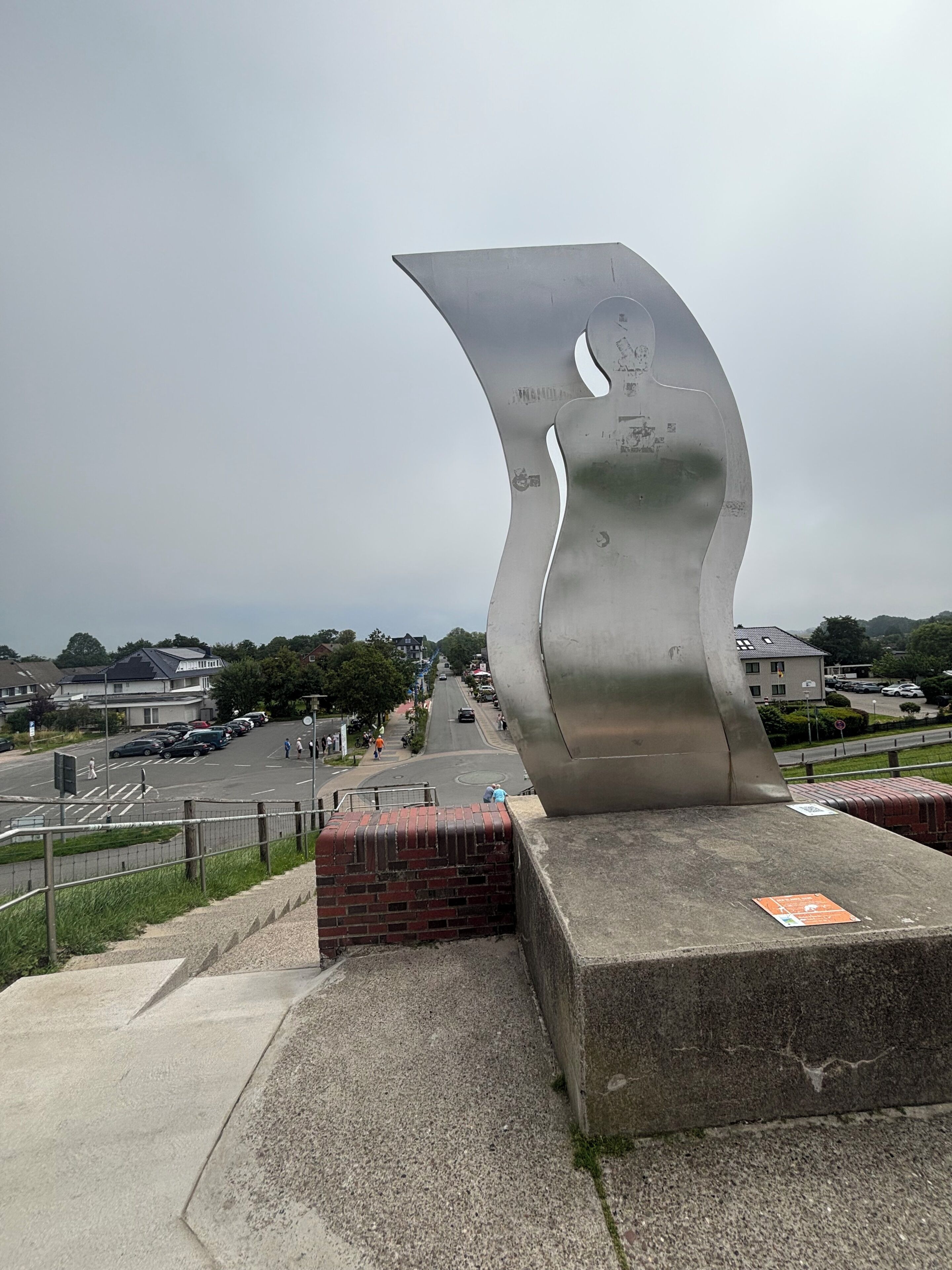 A metal sculpture with a wavy design, located in an urban setting with a road and buildings in the background.