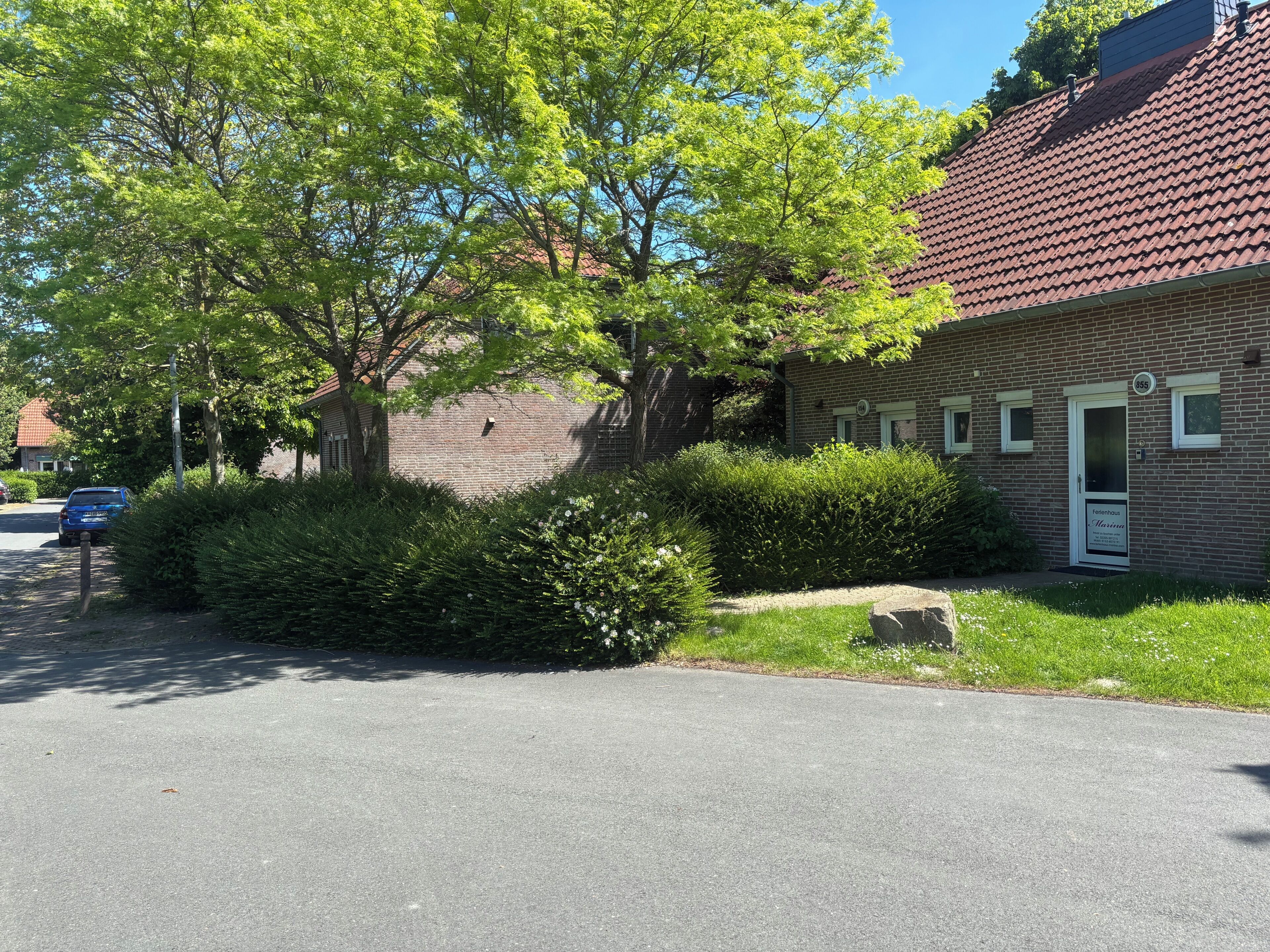 A brick building with a red-tiled roof, a white door, and a small garden area with bushes and a tree.