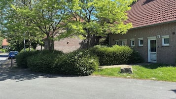 A brick building with a red-tiled roof, a white door, and a small garden area with bushes and a tree.