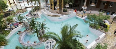 An indoor swimming pool with multiple levels and a central fountain, surrounded by palm trees and lounge areas.