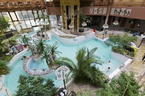 An indoor swimming pool with multiple levels and a central fountain, surrounded by palm trees and lounge areas.