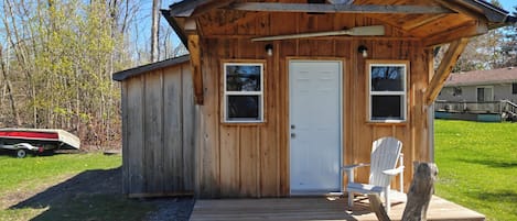 Una cabaña de madera con puerta, dos ventanas, una terraza y una silla.