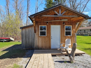 Une cabane en bois avec une porte, deux fenêtres, une terrasse et une chaise.