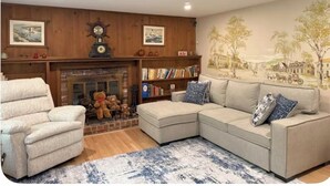 A living room with a fireplace, a sectional sofa, a chair, and a bookshelf. - Grammy’s Cape Cod Summer House (Brewster)