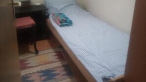 A small, single-bed room with a plain wall, a window, a small table, and a chair.