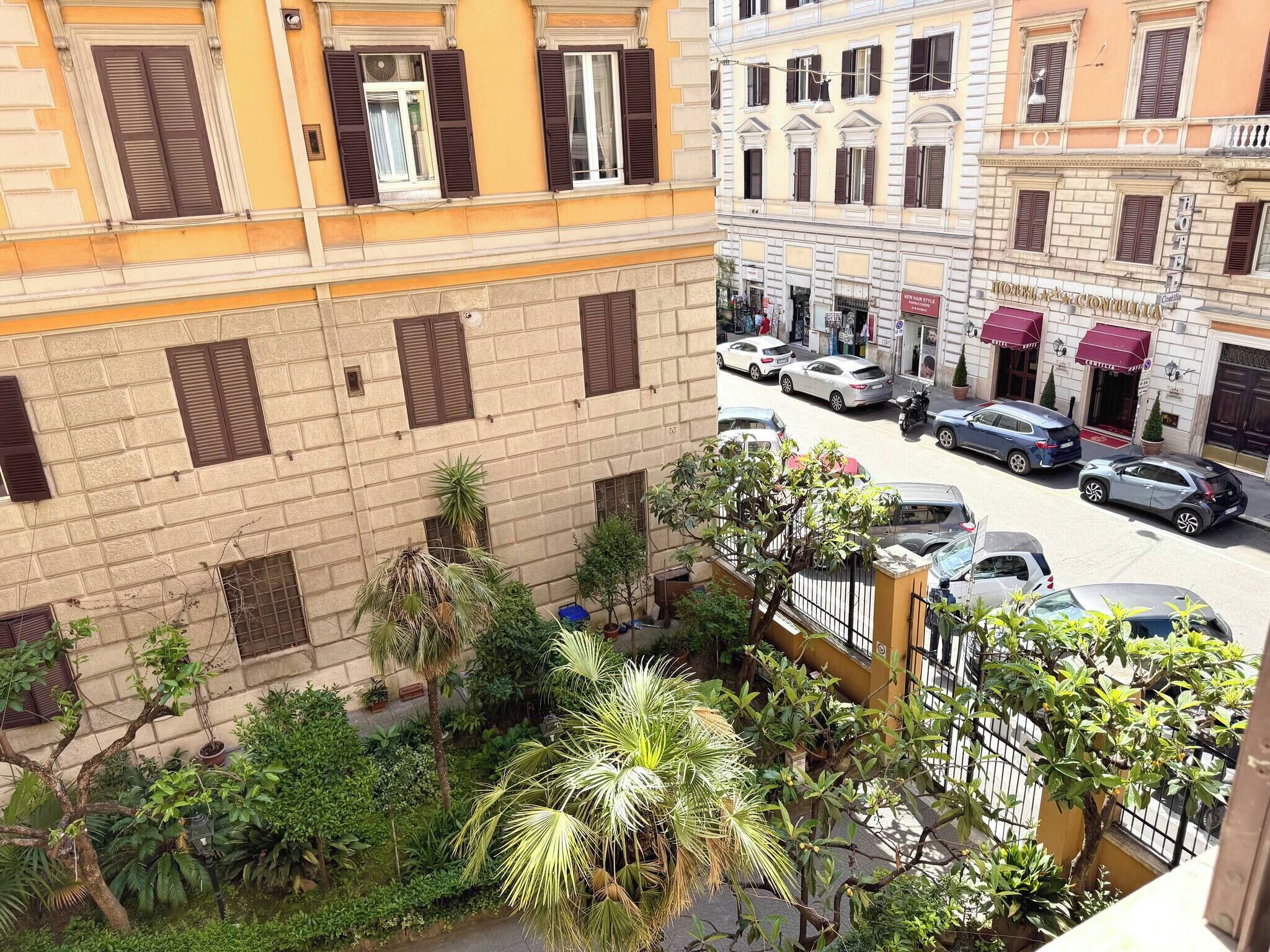 A multi-story building with a courtyard, featuring a variety of plants and a street with parked cars.