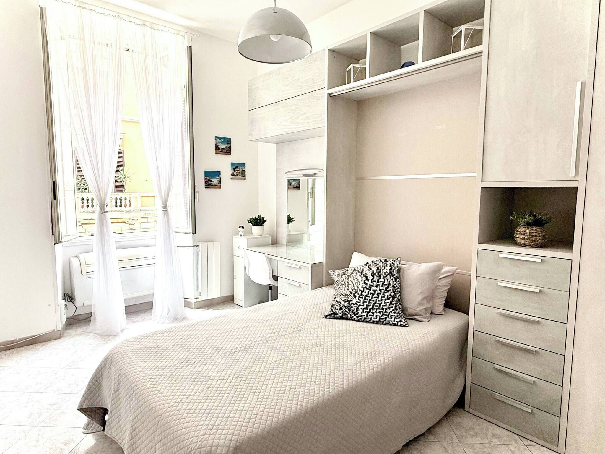A bedroom with a bed, a dresser, a vanity, a mirror, and a window with curtains.