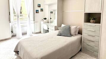 A bedroom with a bed, a dresser, a vanity, a mirror, and a window with curtains.