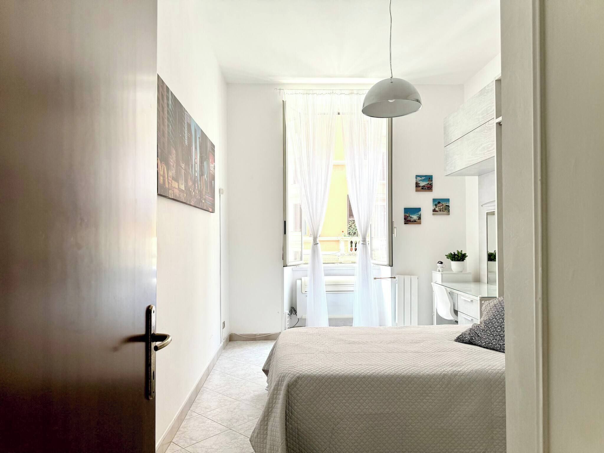 Ein schmaler Flur, der in ein Schlafzimmer mit Bett, Heizkörper, Pendelleuchte und einem Fenster mit durchsichtigen Vorhängen führt.