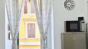 A room with a view through curtains to a building with shutters, a wall clock, and a microwave on a cabinet. - Central Rome Apartment Termini - Roman Haven (Rome)