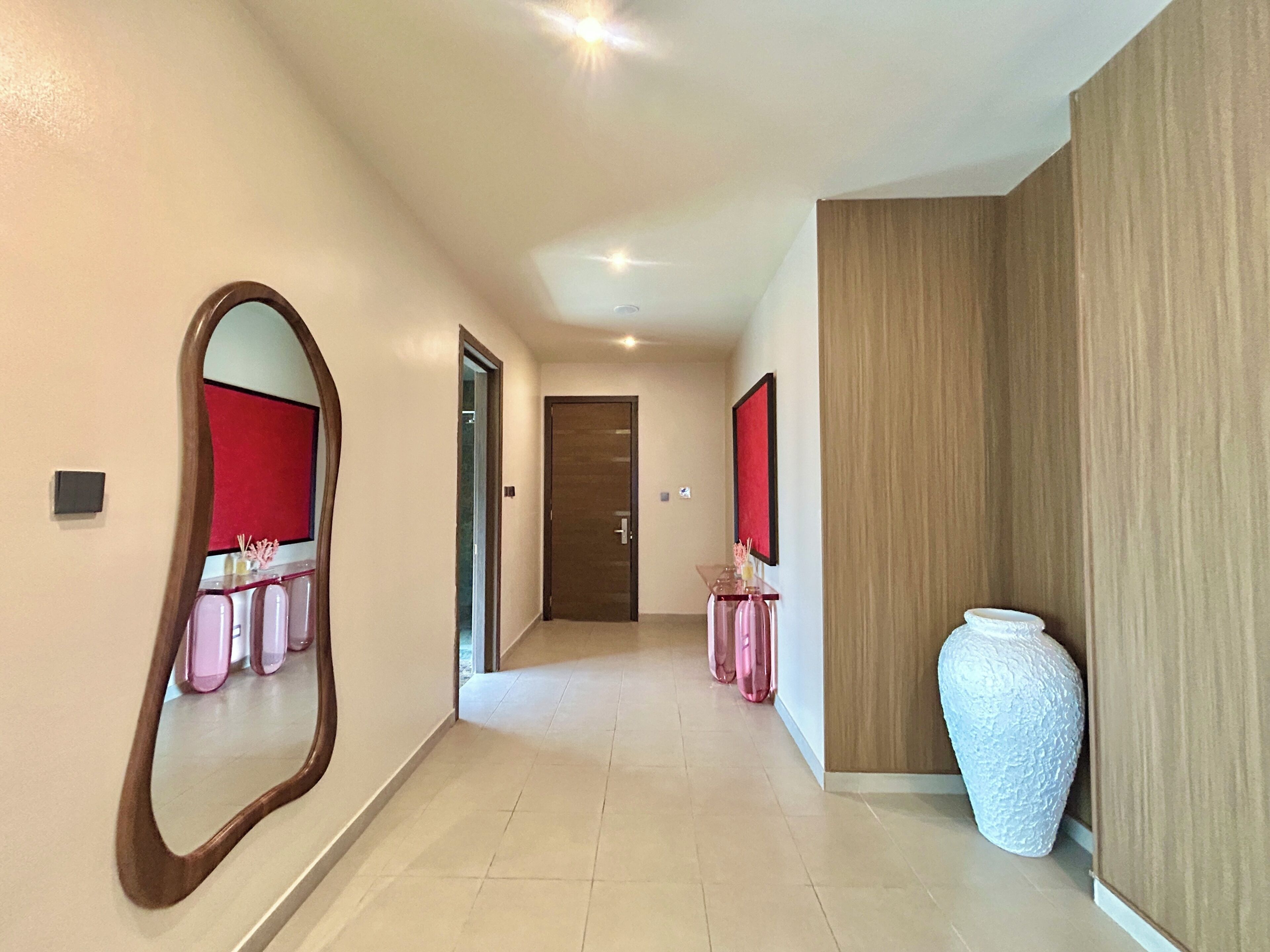 A hallway with a mirror, a door, a console table with pink items, and a white vase.
