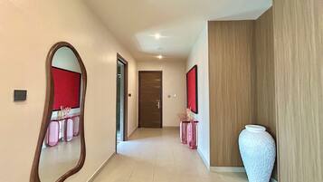 A hallway with a mirror, a door, a console table with pink items, and a white vase.