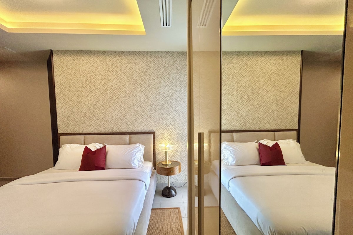Une chambre d’hôtel aux murs recouverts de miroirs, avec deux lits, une table de chevet, une lampe et un mur orné d’un motif.