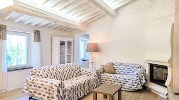 Ein gemütliches Zimmer mit Holzbalkendecke, Kamin, einer Couch, einem kleinen Tisch und einem Fenster mit Vorhängen.